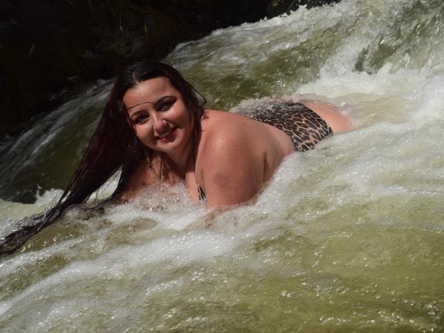 NaomiQuinn met nat, bruin haar, lachend, liggend in een rivier, met een bikini broekje met luipaardprint, haar billen en borsten gedeeltelijk zichtbaar, water spettert om haar heen..