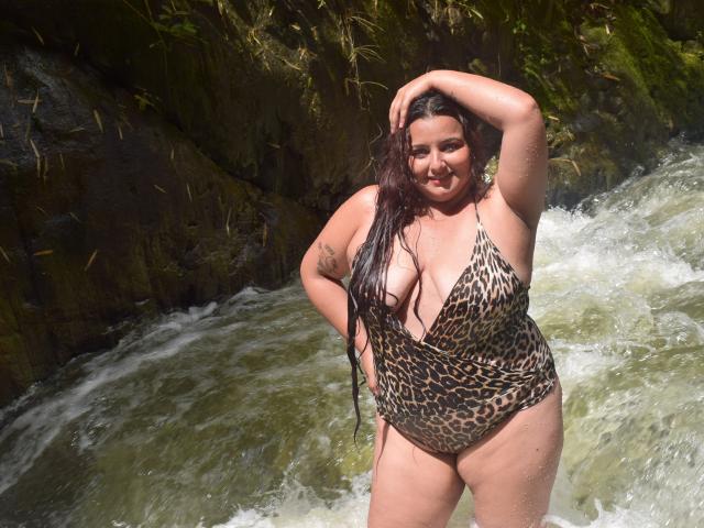 NaomiQuinn, avec ses longs cheveux noirs et mouillés, portant une combinaison une pièce à motifs léopard, se tient dans un ruisseau rocheux, une main sur la tête, souriante, montrant ses seins, avec un tatouage sur son bras..