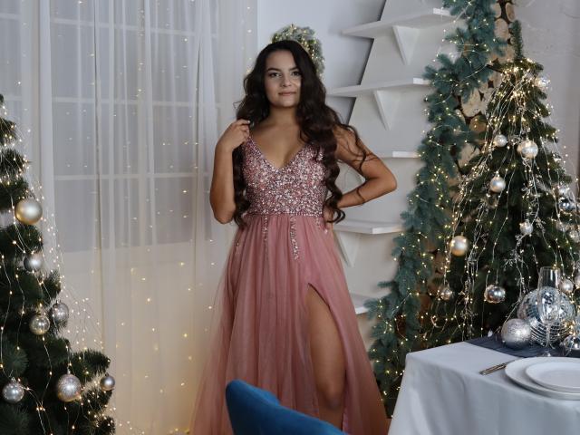 JasmineAsha with long dark hair, wearing a pink, sparkly, deep V, high-slitted dress, standing by decorated Christmas trees, looking confident, with a blue chair in front.