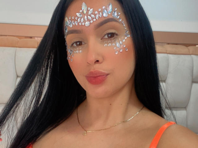 EmilyParkerr has long black hair, light brown skin, and silver leaf-like designs on her face. She's wearing an orange top, necklace, and has red lipstick, smiling softly.