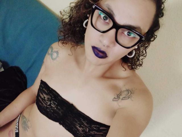 JohndaX with curly brown hair, black lace strapless top, blue lipstick, glasses, ear gauges, tattoos on shoulders, looking up with surprised expression, fair skin, small boobs.