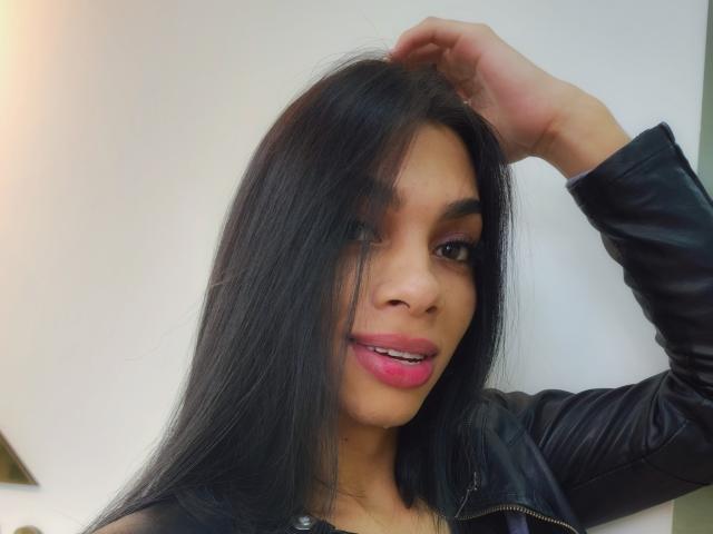 CelesteSweety hat dunkles, glattes Haar, pinken Lippenstift und braune Augen, mit einem leichten Lächeln, und trägt eine schwarze Lederjacke, wobei eine Hand im Haar ist..