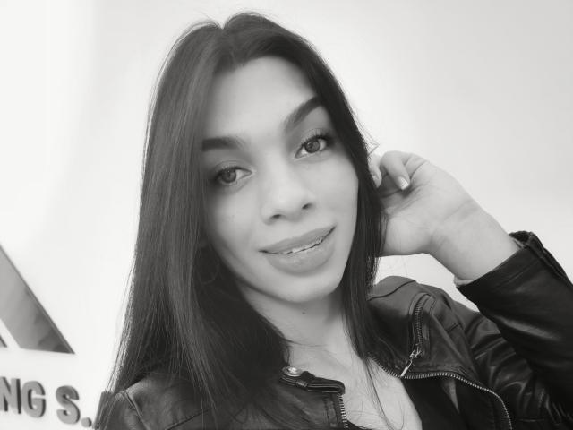 CelesteSweety a de longs cheveux noirs, du rouge à lèvres rose, et porte une veste en cuir noir. Elle a une main dans ses cheveux, et sourit avec la langue légèrement sortie..