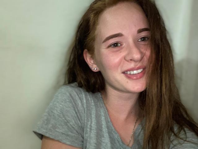 HereIsSoul with long brown hair, gray tee, smiling, showing white teeth, small earrings, and a cute face, looking slightly to the side, with a relaxed, happy expression.