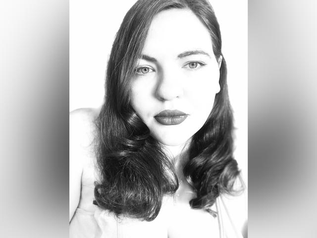 Ein Schwarz-Weiß-Foto von AglayaFame mit welligem, dunklem Haar, vollen Lippen mit dunklem Lippenstift und einem ernsten Gesichtsausdruck. Ihre nackten Schultern sind sichtbar. Sie blickt direkt in die Kamera..