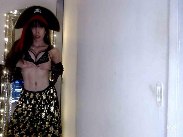 SeleneBrigth with long black hair, pirate hat, black lace bra, and gold-patterned skirt, covering her tits with hands, standing in a doorway, wearing black lace gloves.