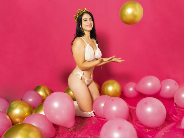 MollyBron kniet inmitten von rosa und goldenen Luftballons, trägt einen weißen Bikini und eine goldene Tiara, hat die Hände ausgestreckt, lächelt und zeigt ihre Brüste und ihren Hintern. Sie hat langes, schwarzes Haar..
