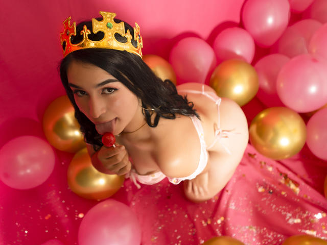 MollyBron com uma coroa dourada, lambendo um pirulito vermelho, ajoelhada em meio a balões rosa e dourados, vestindo lingerie branca que expõe seus seios e nádegas, com cabelos escuros e ondulados..