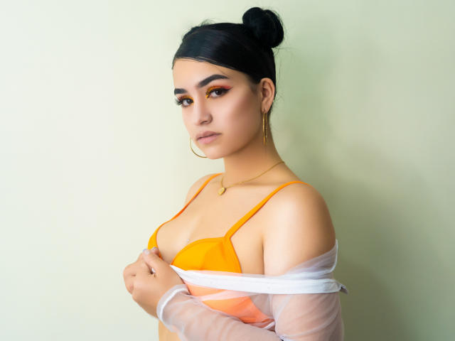 MollyBron, com cabelo escuro preso em um coque, usando um sutiã laranja e uma blusa branca transparente, segurando-a para expor os seios, olhando para a câmera com delineador laranja e brincos de argola dourados..