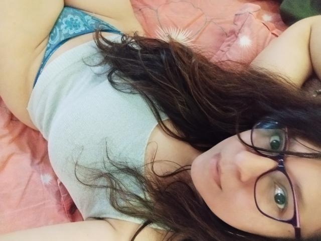 Angelonix with long brown hair, glasses, gray tank top, and blue lace panties, lying on pink bed, showing her tits and pussy, looking at the camera.