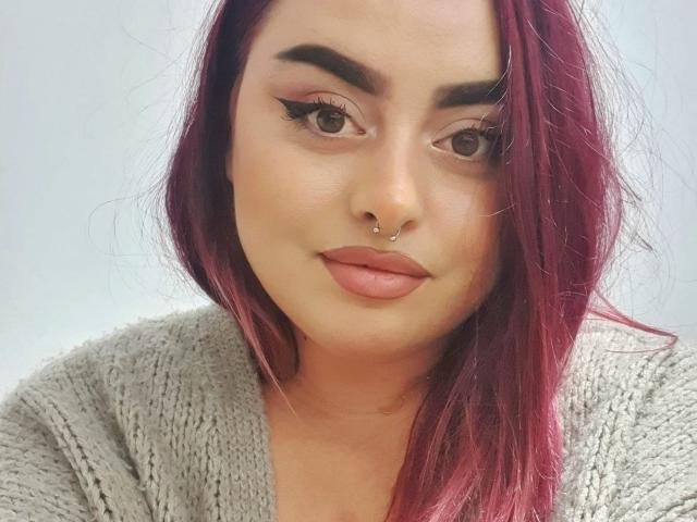 ChaudePourxToi ha i capelli rosso-rosa, un piercing al naso e labbra piene di colore rosa. Indossa un maglione grigio a maglia, con un trucco per gli occhi molto marcato e sopracciglia folte..