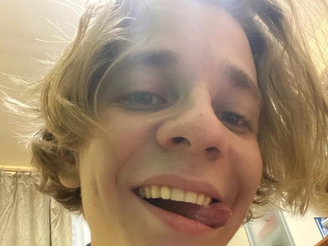 FlexKitty has a goofy smile with tongue out, messy blonde hair, and light skin, showing white teeth. Their tongue is slightly pink, and they have green eyes.