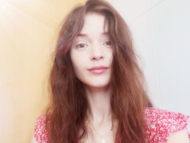 SonyaLovelace with long, messy brown hair, wearing a red, white-patterned top, has pink lips, and a small necklace with a heart pendant, looking softly at the camera.