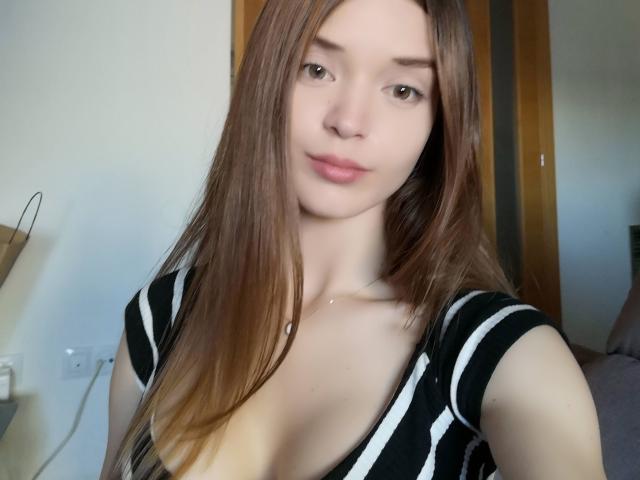SonyaLovelace, com cabelos castanhos longos, vestindo uma blusa listrada preta e branca, que mostra seus pequenos seios e um decote discreto, tem uma expressão neutra e está tirando uma selfie..
