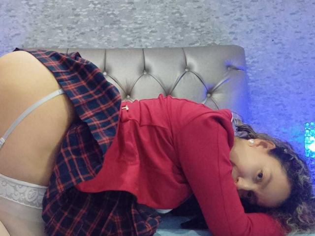 EroticSasha, with curly brown hair, wearing a red shirt and plaid skirt, bends over on a bed, showing her white garter and bare ass. She's looking at the camera.
