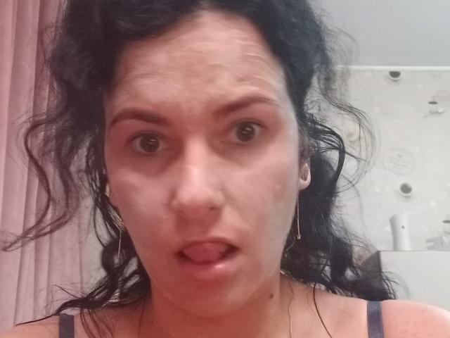 KinkyKosmina with messy black hair, brown eyes, and small earrings, tongue slightly out, looking surprised. She has fair skin and is wearing a dark strap.