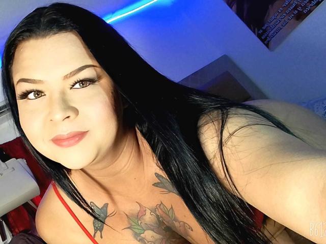 Een selfie van AlexaBigTs met lang zwart haar, bruine ogen en een getatoeëerde borst met vlindertattoos. Ze draagt een rode top en glimlacht licht. Een blauw LED-licht is te zien op de achtergrond..