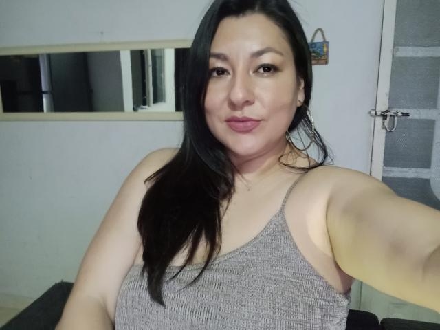 SakotoDika with long black hair, wearing a gray ribbed tank top, showing her tits, puffy pink lips, and a slight smile, taking a selfie in a dim room.