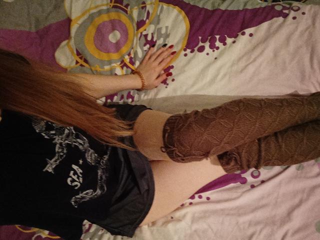 SweetyPieX lying on bed, facing away, wearing black shirt, brown knit leg warmers, showing bare ass, long red hair, red nails, wrist bracelet, purple and yellow patterned bedspread.