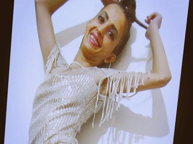 MelaniaTom con un abito lucido, ricoperto di paillettes e con maniche frangiate, sdraiata, braccia alzate, sorridente con labbra rosse, capelli castani raccolti in uno chignon, sfondo bianco, ombra visibile..