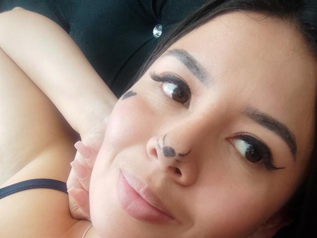 Le visage d'AshleyKatelyn, avec son eye-liner noir, ses tatouages sur le nez et un sourire discret, on peut voir les bretelles de son débardeur noir, elle est allongée et sa main est près de sa joue..