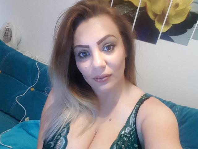 RachelI with blonde-to-brown ombre hair, green eyes, and a mole on her cheek, wearing a green lace top, showing her big tits, sitting on a blue couch.
