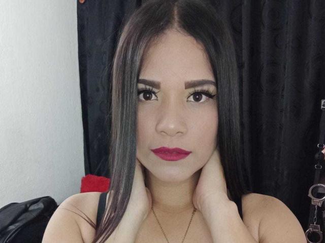 SelenaRogue tem cabelos pretos longos, olhos escuros com cílios grossos, batom vermelho e está tocando o pescoço com as mãos. Ela está vestindo uma blusa preta e olhando diretamente para a câmera..