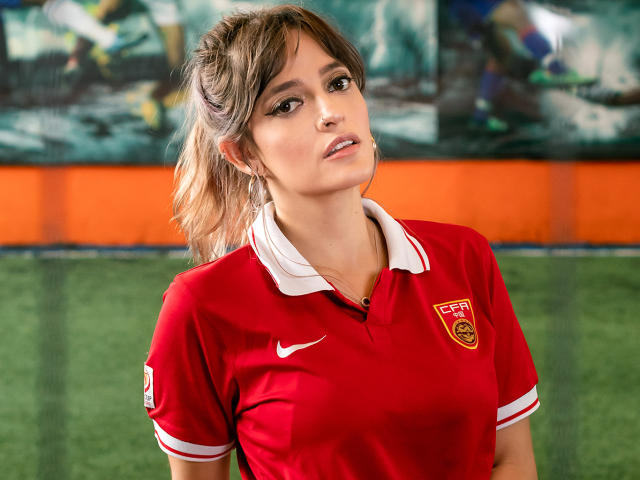 JaneRoa, red soccer jersey with white collar, Nike logo, sports bra, tits visible under jersey, blonde ponytail, green soccer field, serious expression, blurred background, indoor lighting.