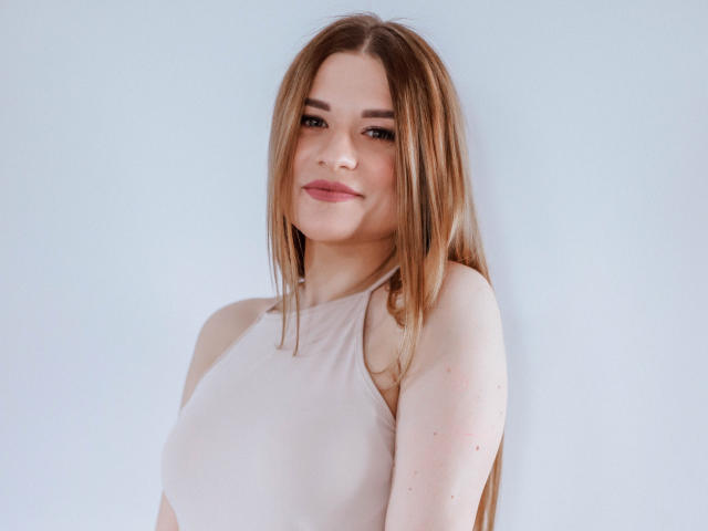 SugarrSanndy with long brown hair, light pink sleeveless top, small tits, subtle smile, and fair skin, standing against a plain light blue background.