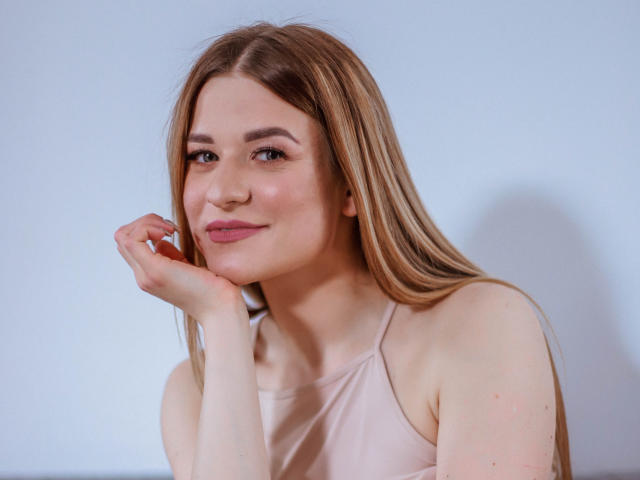 SugarrSanndy heeft lang blond haar, een lichte huid, en draagt een roze tanktop. Ze glimlacht zachtjes met haar hand onder haar kin, en ziet er schattig en ontspannen uit..