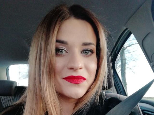 SugarrSanndy hat blondes Haar, grüne Augen und roten Lippenstift. Sie trägt einen schwarzen Sicherheitsgurt im Auto und blickt mit einem leichten Lächeln direkt in die Kamera..