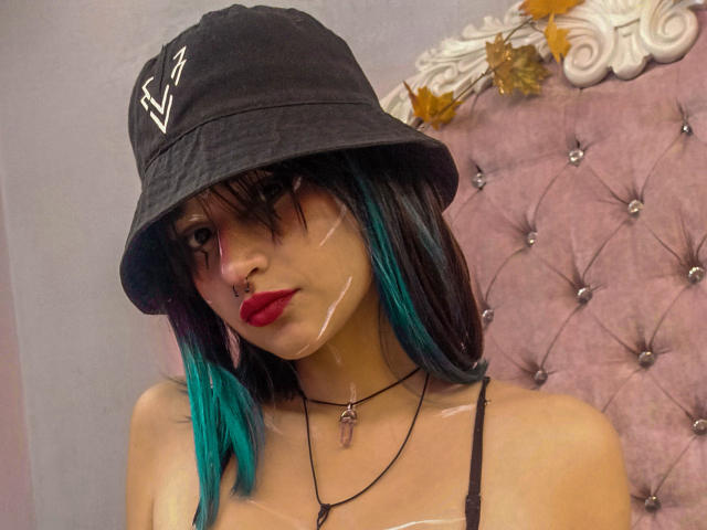 CameroonSweet con cappello a secchiello nero, capelli verde acqua e neri, rossetto rosso, piercing al naso, top nero con spalline, collana con ciondolo a forma di chiave, trucco sul viso e un'aria sensuale, in piedi di fronte a una testiera rosa trapuntata..
