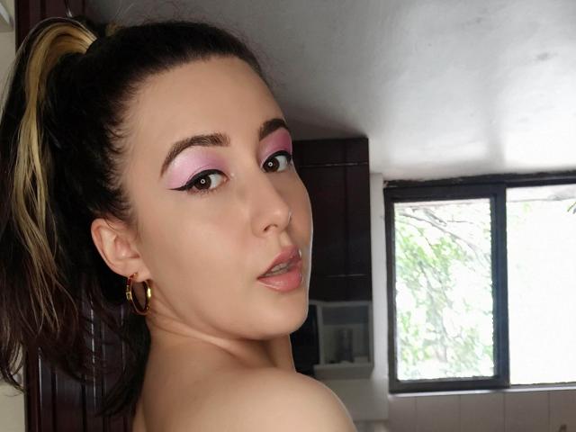 MabyLegrand has pink eyeshadow, a ponytail, gold hoop earrings, and is looking over her shoulder with her tongue slightly out. She's topless with visible shoulders.