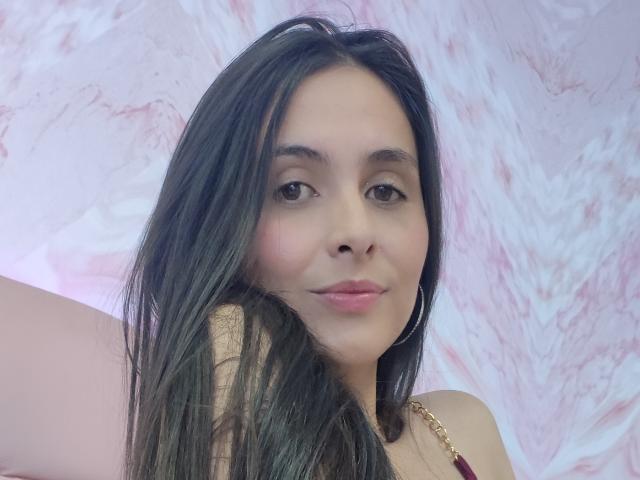AmbeerBlack with long black hair, brown eyes, and pink lipstick, smiling softly, wearing a gold necklace with red beads, holding her hair on the left side.