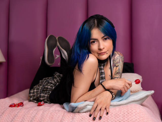 CandiceValo with blue hair, plaid dress, black tank, tattoos, holding red lollipop, lying on pink bed, legs up, sneakers visible, black nails, slightly smiling, cute, sexy.