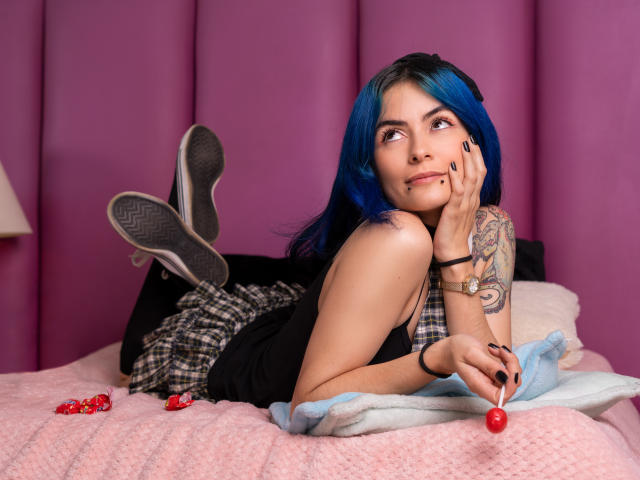 CandiceValo, avec ses cheveux bleus, son débardeur noir, sa jupe à carreaux et ses baskets noires, est allongée sur un lit rose, tenant un bonbon rouge, avec un bras tatoué, des ongles noirs, et un regard mignon et sexy..