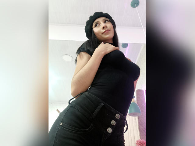 AnyaJay with tan skin, black beanie, and tight black top and pants, showing her large ass with one hand on it, long pink nails, looking down at the camera.