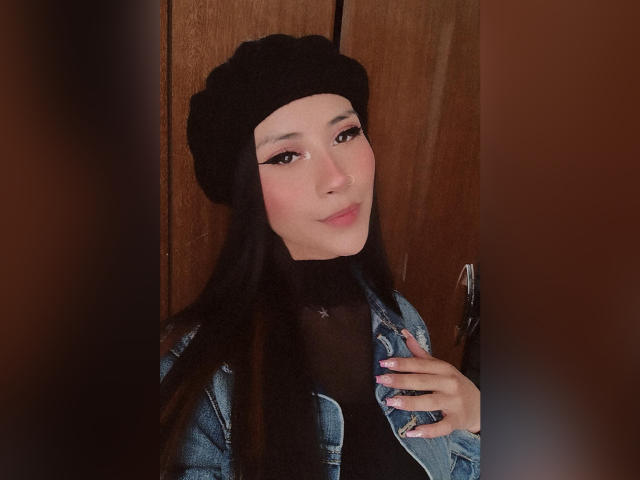 AnyaJay with long black hair, wearing a black beanie and denim jacket with a tear on the right side, looking sideways with a slight smile. Her tits are small.