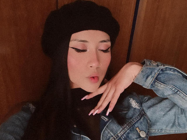 AnyaJay with long black hair, wearing a black beanie, denim jacket, and black turtleneck, smiling, has a cute nose ring, and is touching her chest with manicured nails.