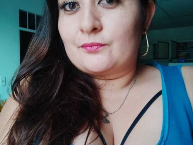 HotValeryPassione a les lèvres roses, les yeux marron, de longs cheveux bruns ondulés, un haut bleu, un soutien-gorge noir à bretelles, des boucles d'oreilles dorées et un petit collier avec un pendentif, montrant ses seins..