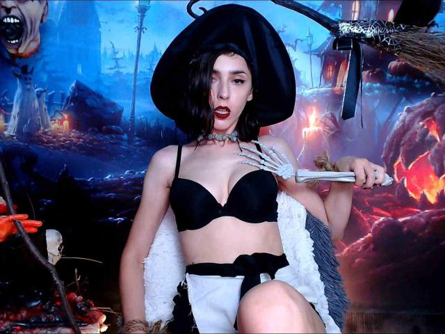 AliceDaelice with dark hair, black hat, black bra, white shorts, choker, holding a broom, red lipstick, tattoos, sitting, medium tits, pale skin, spooky, witchy, fantasy, sexy.