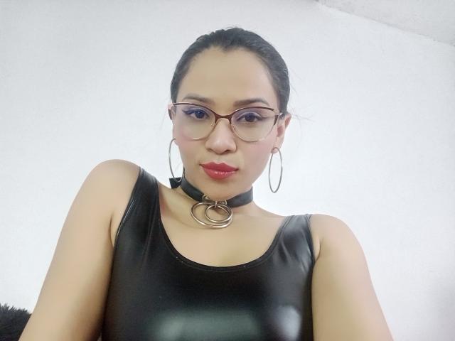 Lily Clark tem cabelo escuro preso em um coque, usa óculos, batom vermelho, um colar preto com um anel, uma blusa de couro preta e brincos de argola. Seus seios são pequenos e ela está olhando para a câmera..