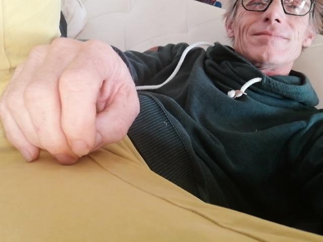 Oboe69 met grijs haar, een bril en een groene hoodie, die met een gesloten rechterhand wijst, draagt een geel shirt, heeft een lichte glimlach en zichtbare rimpels..