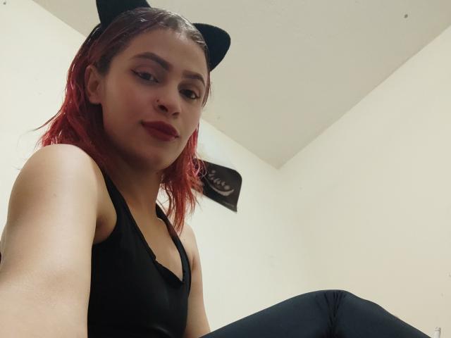 Everlii, com cabelo ruivo, orelhas de gato pretas e uma regata preta, exibindo seus seios em uma selfie tirada de baixo. Ela está sentada com um leve sorriso, vestindo calças pretas..
