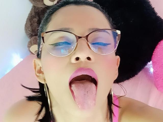 Lily Clark, usando óculos, com sombra azul, batom rosa, língua de fora, cabelo preto, brincos, blusa rosa, brinquedos de pelúcia ao fundo, olhando para cima, close-up do rosto..