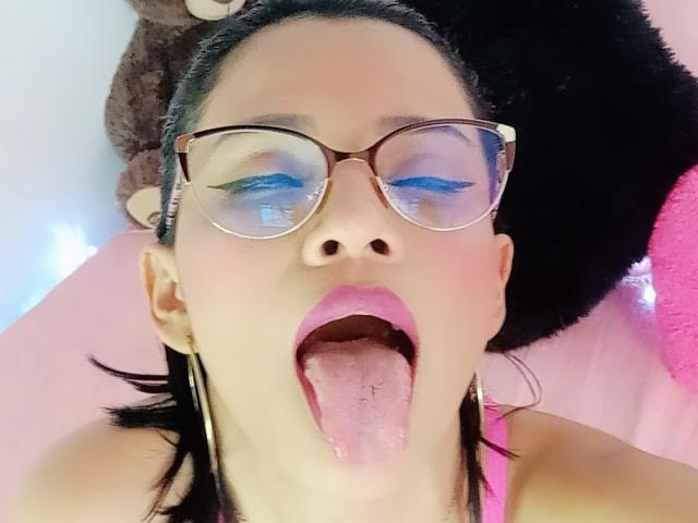 Het gezicht van Lily Clark, met haar tong uit, blauwe oogschaduw, roze lippenstift en bruine bril, zwart haar, gekleed in een roze top, met een pluizig zwart en bruin speeltje achter haar..