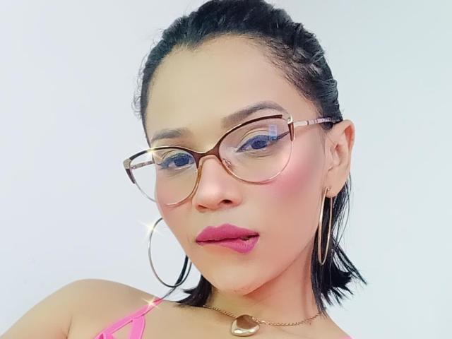 Lily Clark hat rosafarbenen Lippenstift, blaue Augen und trägt goldfarbene Brillen mit großen Ohrringen. Ihr schwarzes Haar ist zurückgekämmt, und sie trägt ein pinkes Oberteil..