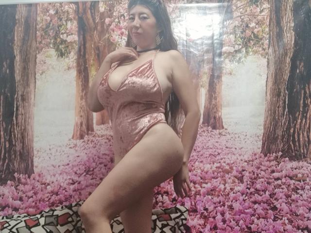 HotPamelaSex with big tits, pink velvet one-piece, black choker, long brown hair, pouty face, standing, right leg raised, pink floral background, patterned rug, soft indoor light.