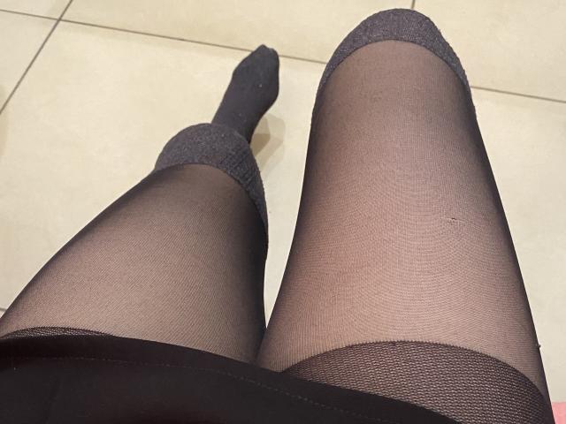 Le gambe di Rubbi, indossanti calze nere trasparenti con parte superiore grigia, e che mostrano i suoi calzini neri senza punta, sono riprese da un'angolazione bassa. Le sue gambe sono leggermente divaricate, evidenziando la texture..