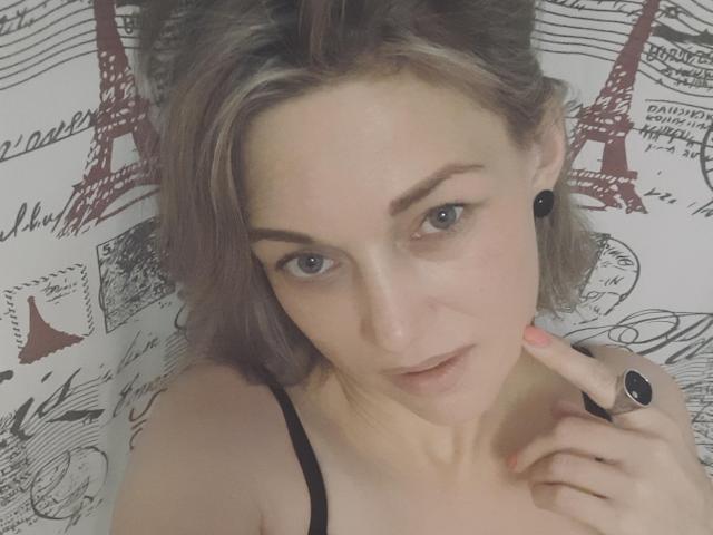 CarmenSun, com cabelo castanho claro, olhos azuis, fones de ouvido pretos, alça preta, dedo tocando o queixo, maquiagem discreta, boca ligeiramente aberta, olhando para a câmera, usando um anel preto..